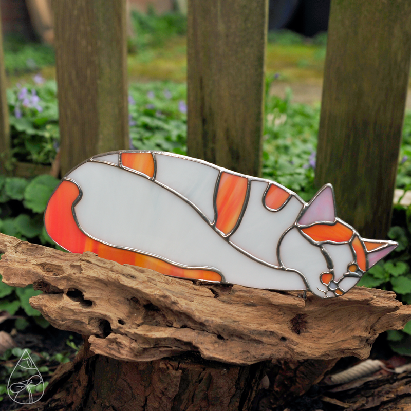 A stained glass piece of a sleeping white and orange cat, mounted on driftwood.