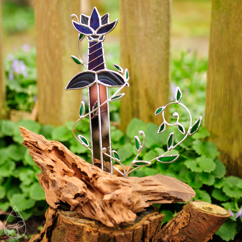 A stained glass piece of a lotus sword imbedded in driftwood, with vines wrapped around the sword.
