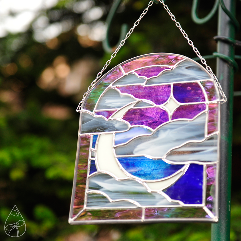 A stained glass suncatcher of a Moonlit Window - featuring a crescent moon, star, and clouds framed in an arched window. 