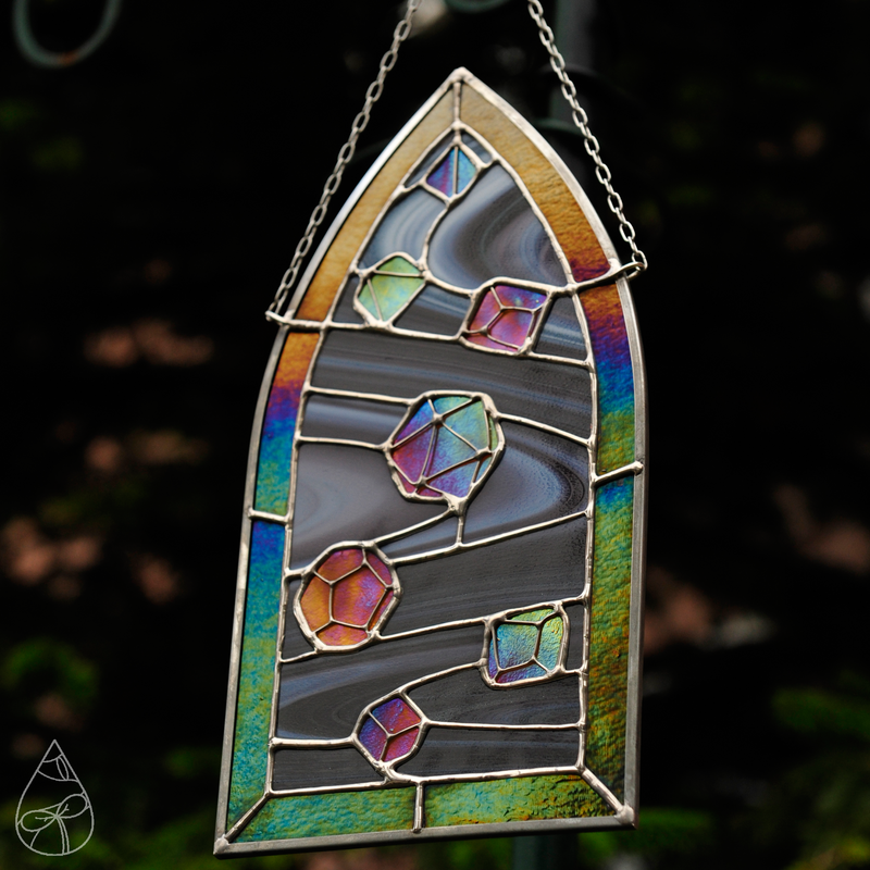 A stained glass suncatcher of iridescent dice tumbling in a swirled background, framed in a gothic shaped window.